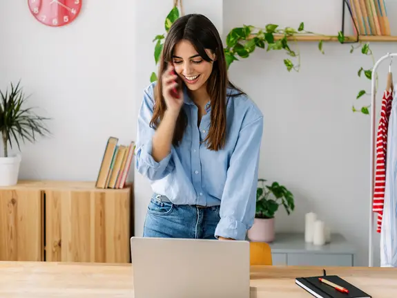 Happy young adult small business female owner talking on mobile phone while working on laptop computer in clothing store